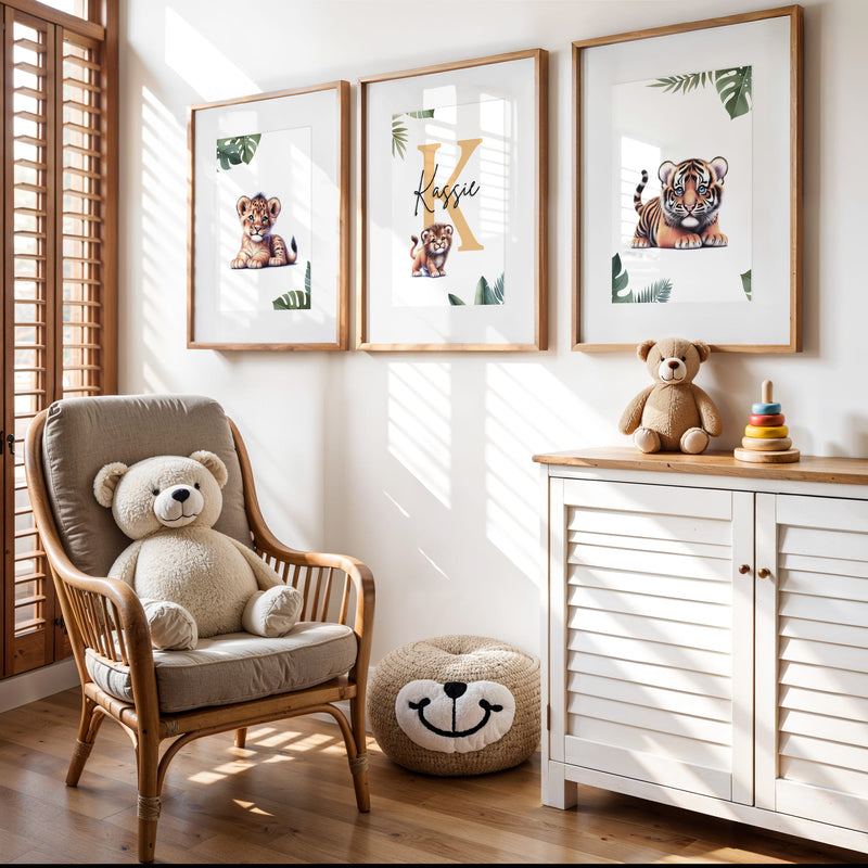 Set of three A4 prints featuring baby big cats - lion cub, tiger cub, and cheetah cub. The animals are depicted in a cartoony, bright style, resembling coloured pencil drawings. Jungle-style leaves adorn the edges of the prints. The middle print features a smaller animal compared to the others, with a large initial letter in the background. A personalised name in black overlays the initial letter.