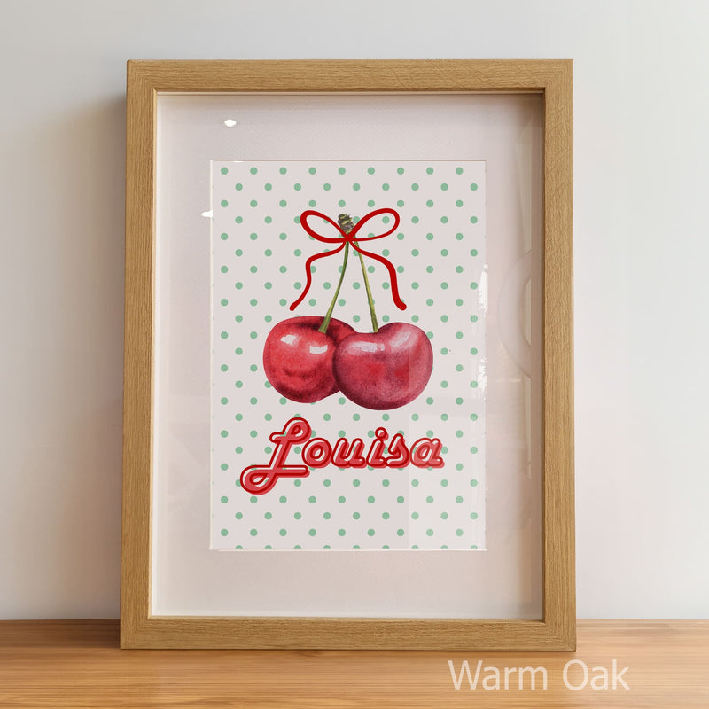Framed artwork with cherries and the name 'Louisa' on a polka dot background, displayed against a light wall.
