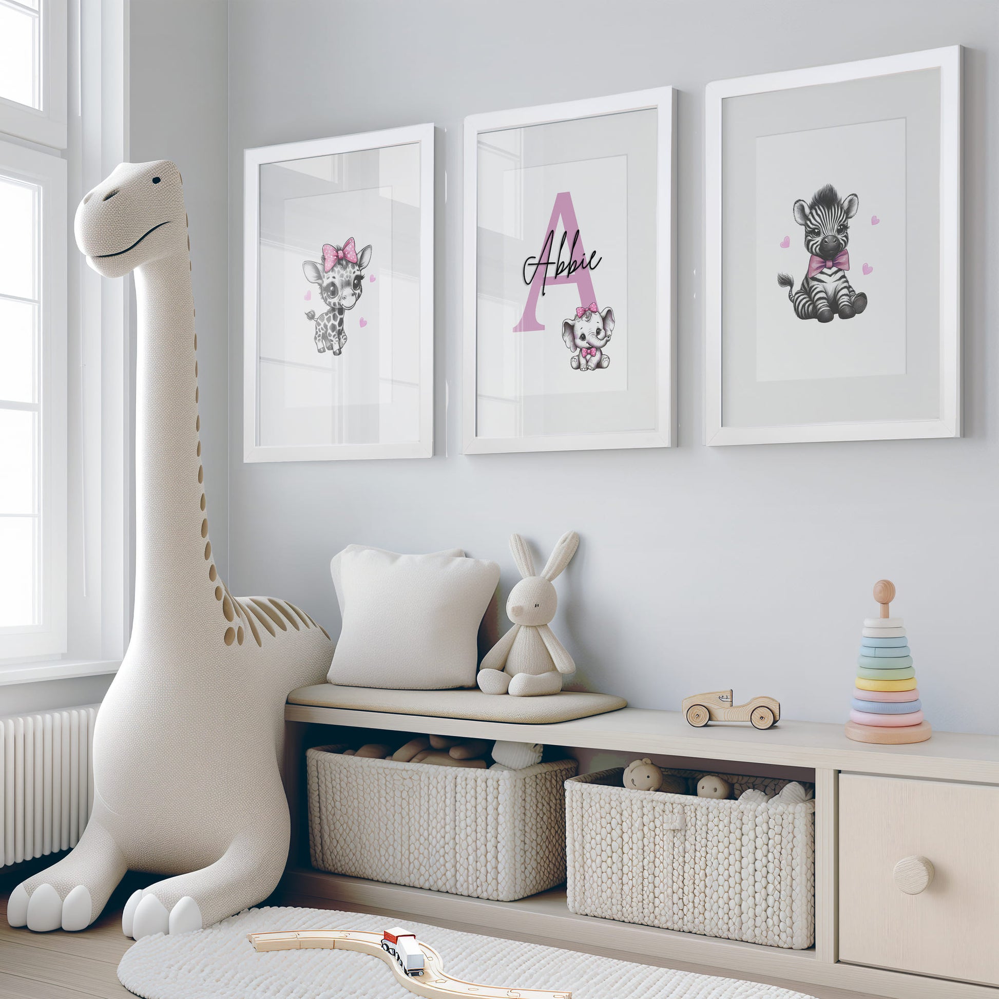 Set of three A4 prints in the style of pencil drawings. Each print features a cartoon baby giraffe, baby elephant, and baby zebra in black and white. They all wear a pink bow, with a few small pink hearts in the background. The middle image contains the elephant, which is smaller than the other two images. In the background, there is a large initial with a personalised name in black.