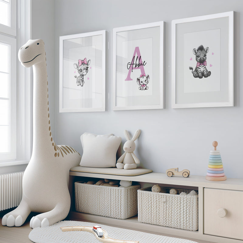 Set of three A4 prints in the style of pencil drawings. Each print features a cartoon baby giraffe, baby elephant, and baby zebra in black and white. They all wear a pink bow, with a few small pink hearts in the background. The middle image contains the elephant, which is smaller than the other two images. In the background, there is a large initial with a personalised name in black.