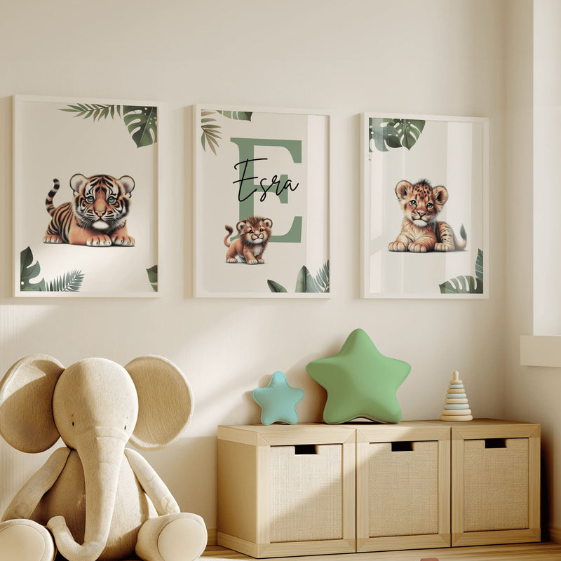 Set of three A4 prints featuring baby big cats - lion cub, tiger cub, and cheetah cub. The animals are depicted in a cartoony, bright style, resembling coloured pencil drawings. Jungle-style leaves adorn the edges of the prints. The middle print features a smaller animal compared to the others, with a large initial letter in the background. A personalised name in black overlays the initial letter.