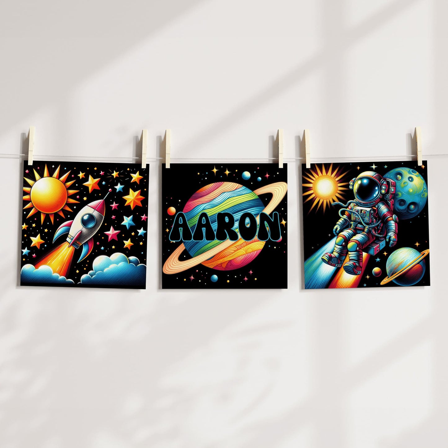 Set of Three Square Nursery Prints. Black background with bright colourful images with a space theme - astronaut, rocket, planet. One print is personalised with a name