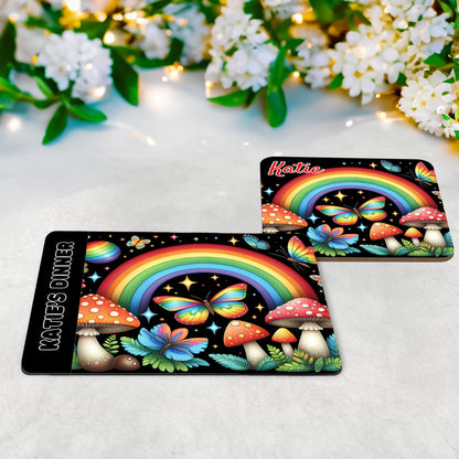 Rainbow coloured bright and bold fairy garden placemat and coaster set. Featuring rainbows, butterflies and toadstools, this multicoloured design pops on its black background, and is personalised with name of your choice
