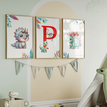 Set of three A4 prints featuring cute baby dinosaurs. The dinosaurs are depicted in a cute, cartoony, bright style resembling coloured pencil drawings. Rainbow coloured leaves decorate the edges of the prints. The middle print features a smaller dinosaur than the others, with a large initial letter in the background. A personalised name in black overlays the initial letter.