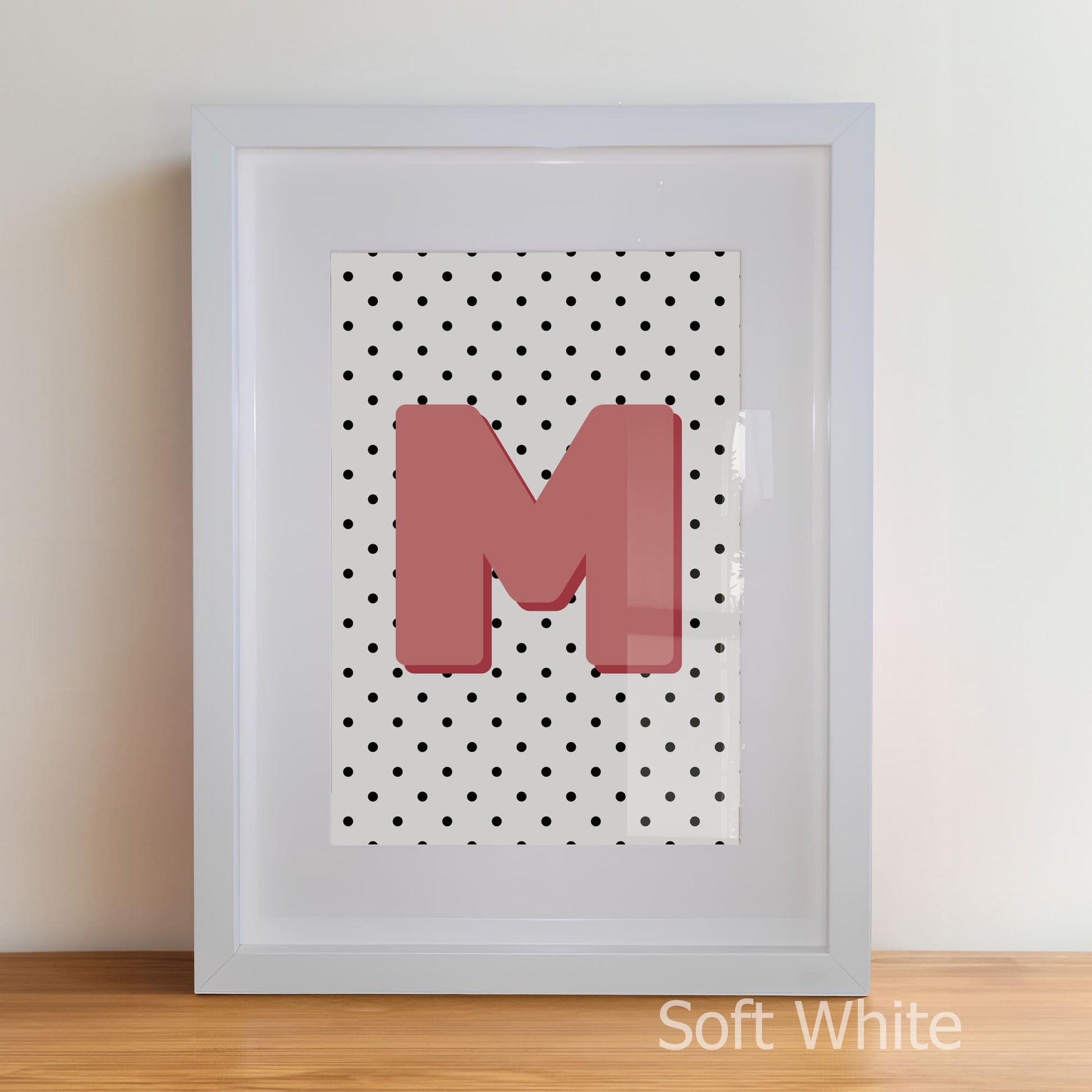 Framed artwork with a pink letter 'M' on a polka dot background, soft white frame.