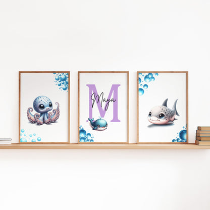 Set of three A4 prints featuring baby ocean animals - whale, shark, and octopus. The animals are depicted in a cute, cartoony, bright style resembling coloured pencil drawings. Blue bubbles decorate the edges of the prints. The middle print features a smaller animal than the others, with a large initial letter in the background. A personalised name in black overlays the initial letter.