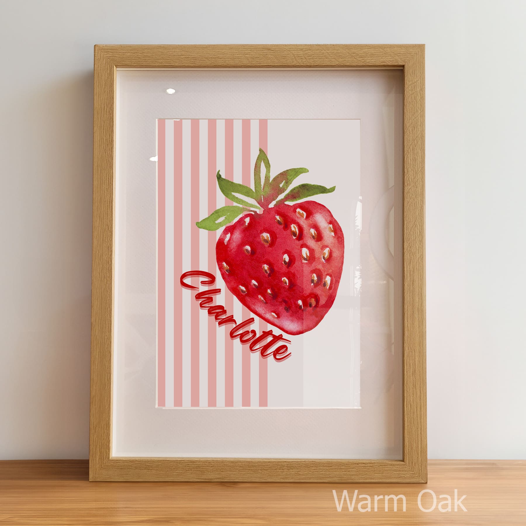 Framed artwork of a red strawberry with green leaves on a striped background, displayed against a light wall.