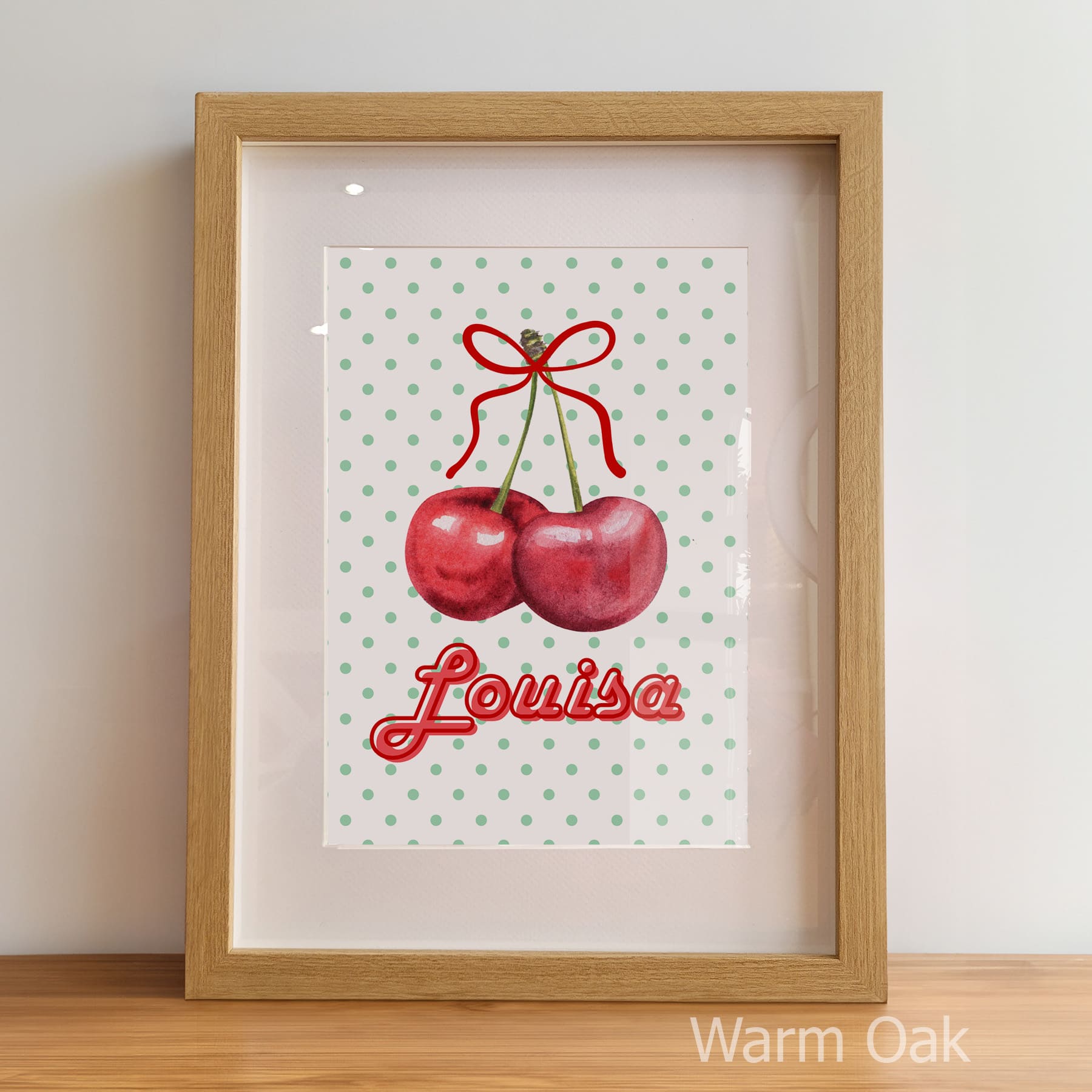 Framed artwork with cherries and the name 'Louisa' on a polka dot background, displayed against a light wall.