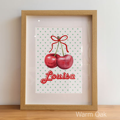 Framed artwork with cherries and the name 'Louisa' on a polka dot background, displayed against a light wall.
