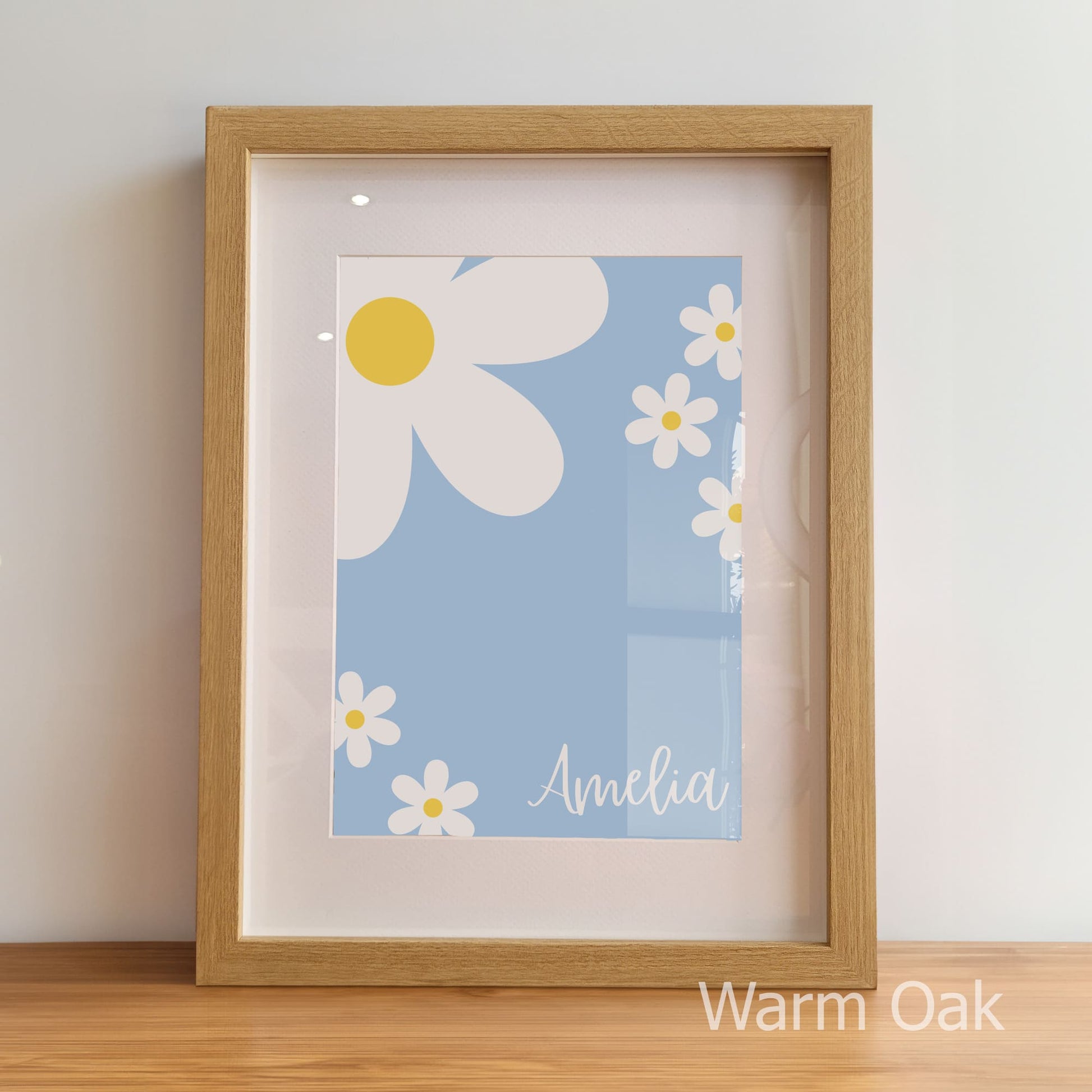 Framed artwork with daisy design and 'Amelia' text on a light blue background, displayed on a wooden surface.