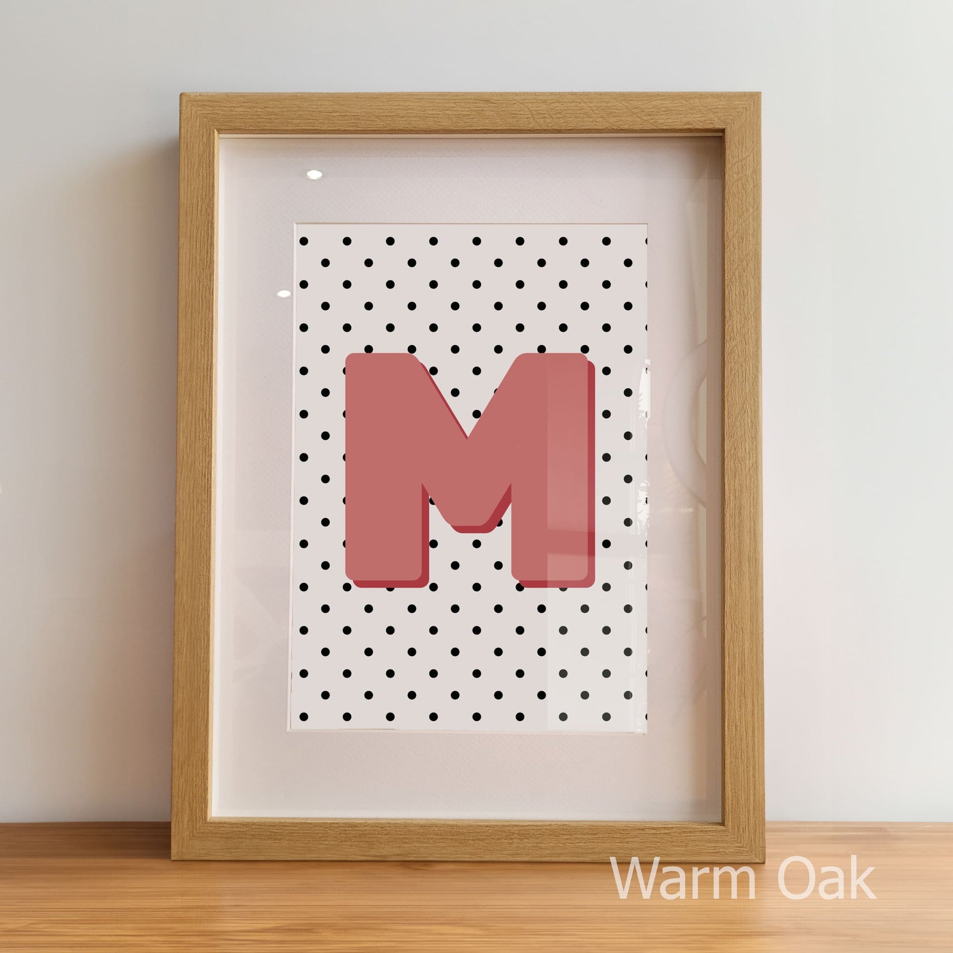 Framed artwork with a pink letter 'M' on a polka dot background, displayed against a light wall.