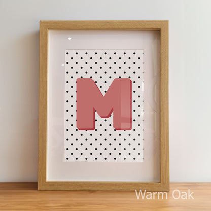 Framed artwork with a pink letter 'M' on a polka dot background, displayed against a light wall.