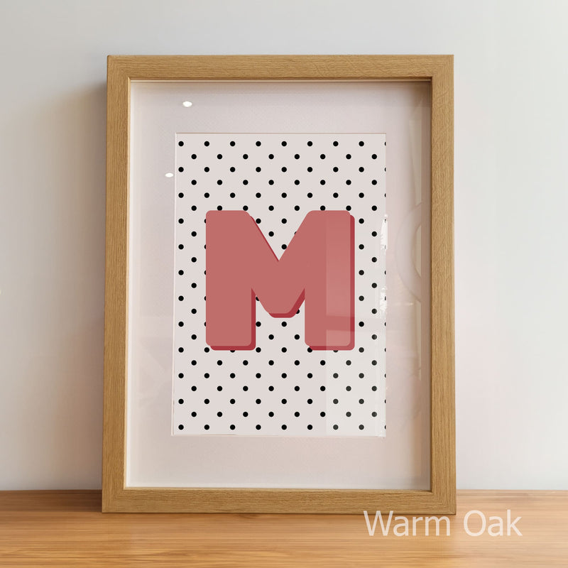 Framed artwork with a pink letter 'M' on a polka dot background, displayed against a light wall.