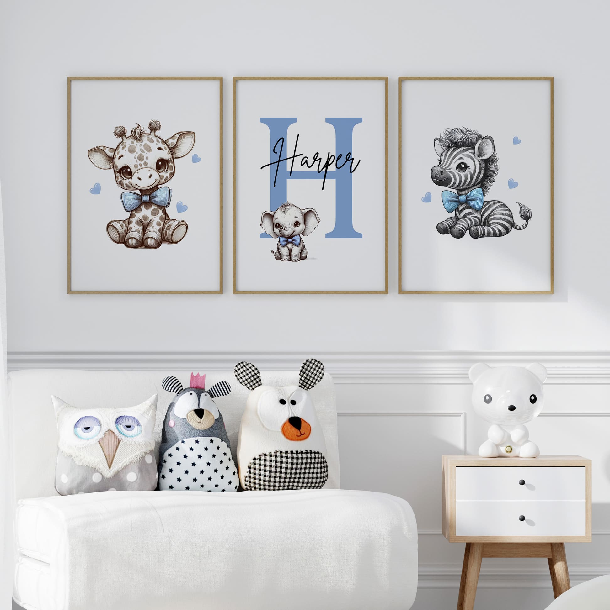 Set of three A4 prints in the style of pencil drawings. Each print features a cartoon baby giraffe, baby elephant, and baby zebra in black and white. They all wear a blue bow, with a few small blue hearts in the background. The middle image contains the elephant, which is smaller than the other two images. In the background, there is a large initial with a personalised name in black.